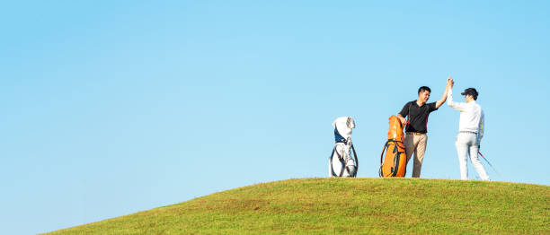 Two golfers high-fiving atop a hill with three golf bags nearby under clear blue sky.
