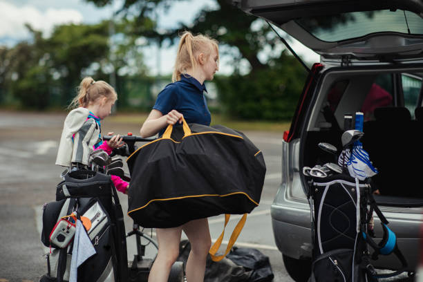 Woman and child loading gear into car trunk; woman carries a black golf bag with yellow trim.
