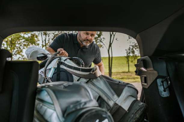 Man loading a grey golf bag into a car trunk, preparing for a game.