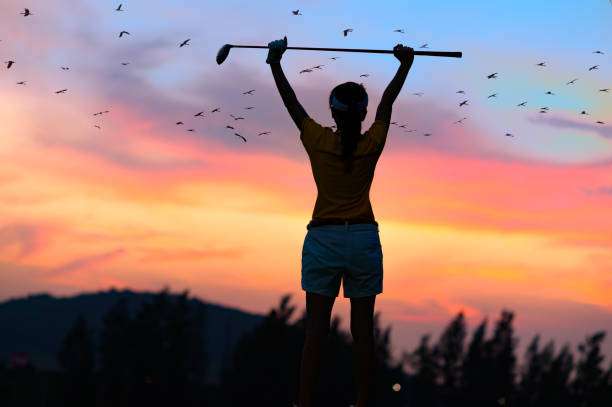 Golfer celebrates with club at sunset, anticipating her next shot with a trusty golf bag.