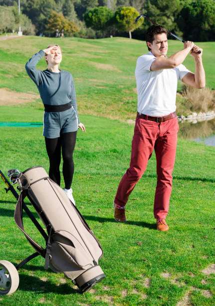 Man swings golf club as woman watches, a golf bag resting nearby on the green.