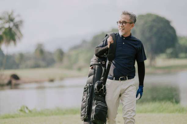 Asian man walking on a sunny golf course, carrying a black golf bag over his shoulder.