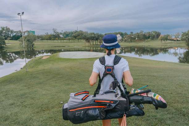 Golfer carries a stand golf bag with clubs on her back, walking toward the green.