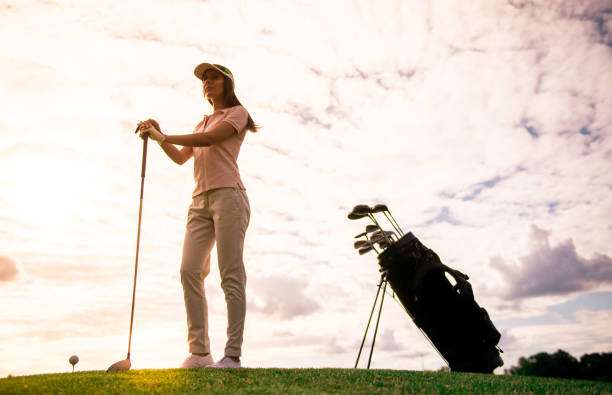 Female golfer with club stands by her Golf Bag, silhouetted against sunset sky.