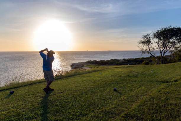 Golfer in silhouette swings on a scenic oceanfront course at sunset, near a golf bag.